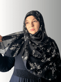 Woman wearing a black chiffon hijab with ivory floral patterns, holding the fabric to showcase its softness and drape.