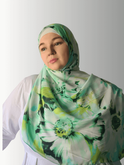 A woman wearing the Spring Dew Chiffon Hijab, featuring a soft green floral design on a white base. The lightweight chiffon drapes elegantly, complementing her modest outfit.
