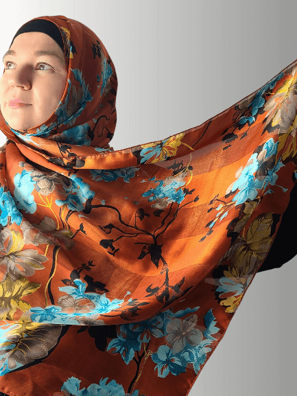 Woman wearing a silk-blend hijab in warm golden orange with blue floral patterns, holding the fabric to display its luxurious drape.