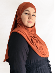 Woman wearing a rich terracotta-orange Instant Jersey Shawl with a matching undercap, demonstrating the lightweight drape and coverage of the fabric against a plain white background.