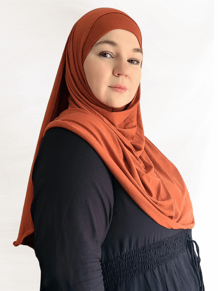 Woman wearing a rich terracotta-orange Instant Jersey Shawl with a matching undercap, demonstrating the lightweight drape and coverage of the fabric against a plain white background.