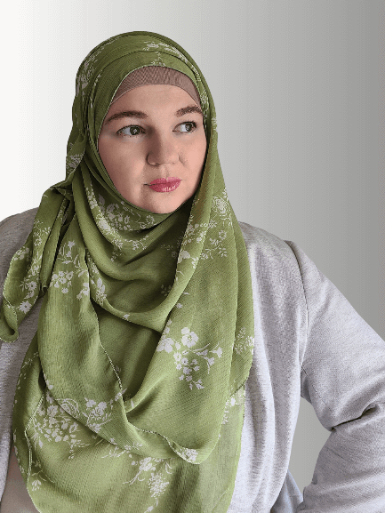 A woman wears an olive green chiffon hijab with a soft floral pattern, draped elegantly.