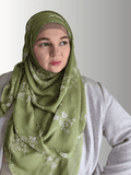 A woman wears an olive green chiffon hijab with a soft floral pattern, draped elegantly.